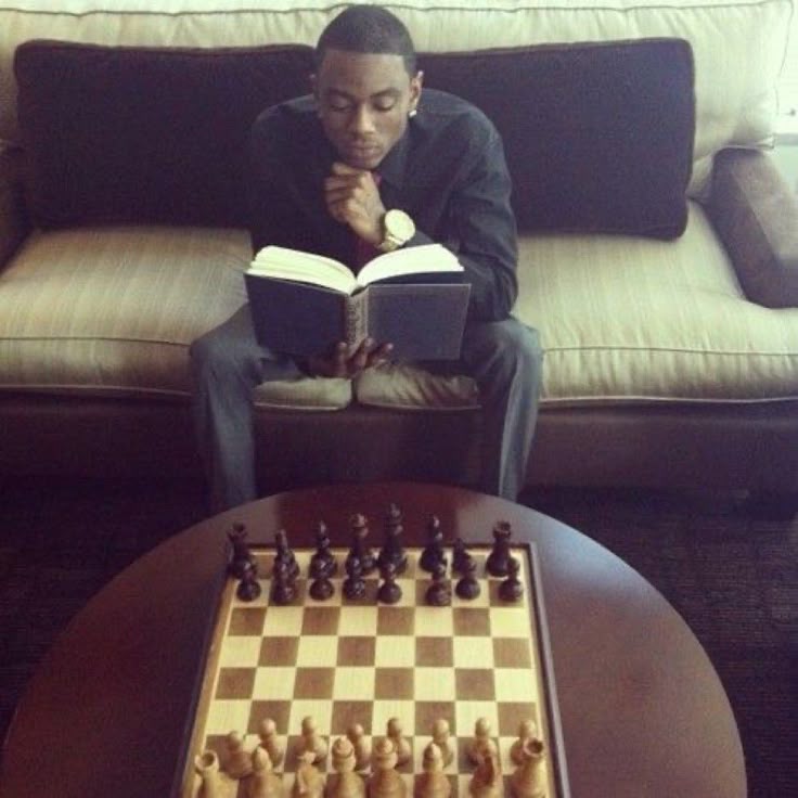 This may contain: a man sitting on a couch reading a book next to a chess board and table