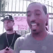 a man is holding a cell phone in front of a fence while another man looks on .