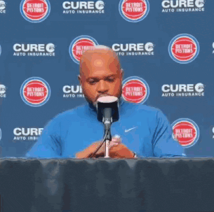 a man in a blue shirt is sitting at a table with a microphone in front of a detroit pepsi sign