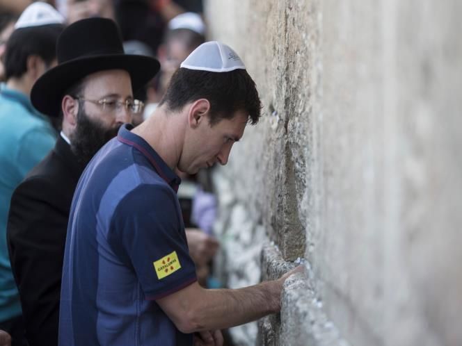 This may contain: a man wearing a blue shirt and black hat standing next to a stone wall with writing on it