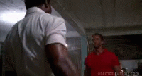 two men are standing next to each other in a room . one of the men is wearing a red shirt .