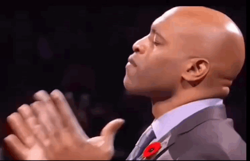 a bald man in a suit and tie with a poppy on his lapel is clapping his hands .