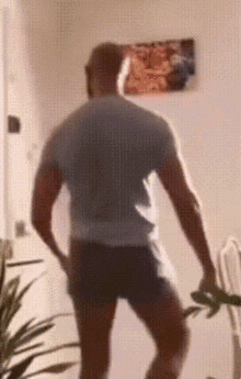 a man in a gray shirt and black shorts is standing in a room with a picture on the wall behind him .