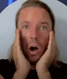 a man with long hair and a beard is making a surprised face while holding his hands to his face .