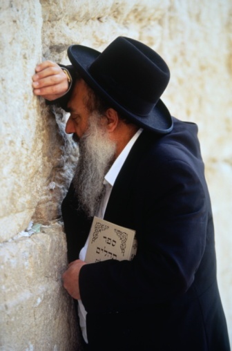 This may contain: a man with a long white beard wearing a black hat and jacket leaning against a stone wall