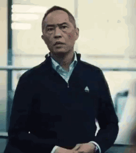 a man in a black jacket and white shirt is standing in front of a window and making a funny face .