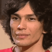 a close up of a man 's face with a red shirt on and a necklace around his neck .