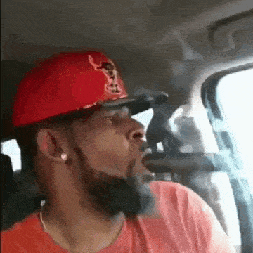 a man with a beard wearing a red hat is smoking a pipe in a car .