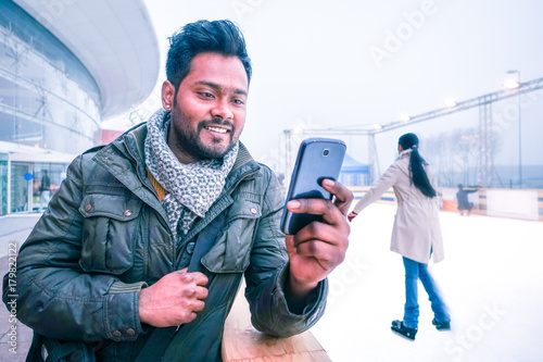 Happy indian man looking mobile phone outdoors at winter with ice skating  scene background - Cheerful bangladeshi young guy using cellphone smiling  looking telephone screen outside on grey light day Stock Photo |