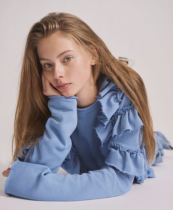 This may contain: a young woman sitting on the floor with her hand under her chin and looking at the camera