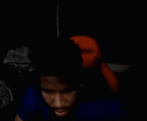 a man in a blue shirt is sitting in a dark room with a stuffed animal behind him .