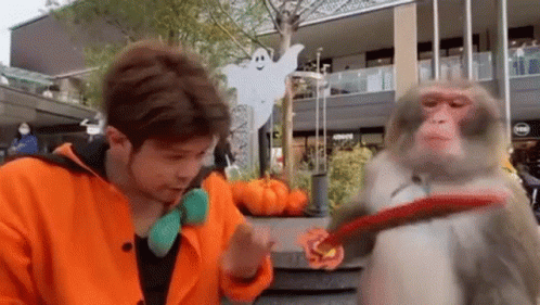 a man in an orange jacket is playing with a monkey on a leash in front of a building .