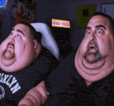two fat men are sitting in front of an arcade machine and one of them is wearing a shirt that says brooklyn