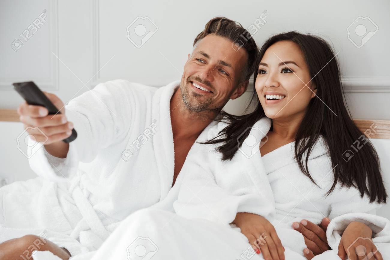 104557929-multiethnic-couple-caucasian-man-and-asian-woman-wearing-white-housecoat-lying-in-home-bedroom-or.jpg