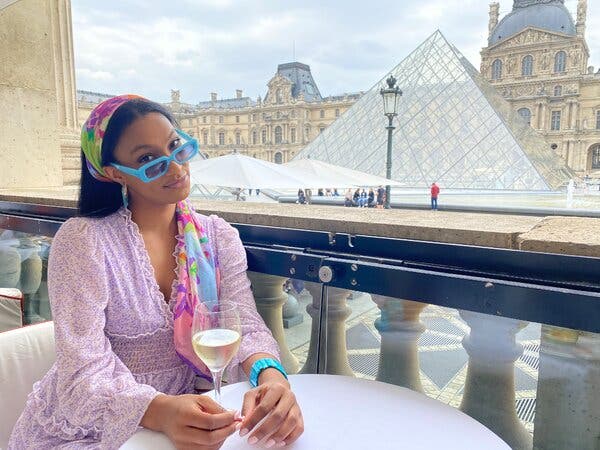A woman in a purple dress and a colorful head scarf poses in front of the Louvre Museum with a glass of champagne.